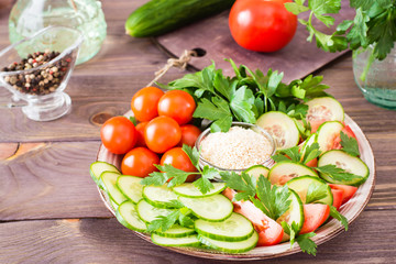 Fresh slices of cucumbers, tomatoes, sesame seeds in a bowl and parsley leaves on a plate on a wooden table. Oil and seasoning for cooking vegetable salad