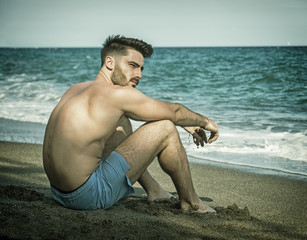 Athletic man at the seaside using cell phone to call someone with the sea behind him, sitting on a rock