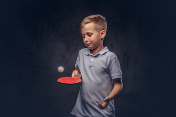 Cute redhead boy dressed in a white t-shirt playing ping-pong in a studio. Isolated on dark textured background. - obrazy, fototapety, plakaty