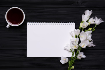 White flowers bells, coffee and empty notebook for your text on a black wooden background.