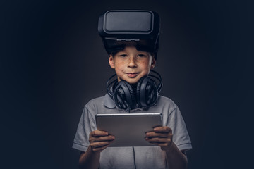 Portrait of a cute little boy dressed in a white t-shirt, with virtual reality glasses and headphones is holding a digital tablet at a studio, isolated on dark background. Education and technology