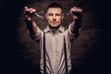 Portrait of a old-fashioned tattooed hairdresser wearing a white shirt with suspenders holds a scissors. Isolated on dark background.