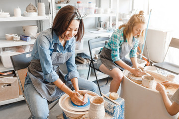 Two girls friends smiling and talking while working on potters wheel making clay handmade craft in pottery workshop, friendship and guidance concept - obrazy, fototapety, plakaty