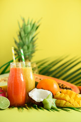 Juicy papaya and pineapple, mango, orange fruit smoothie in two jars on yellow background. Detox, summer diet food, vegan concept. Copy space. Fresh juice in glass bottles over green palm leaves.