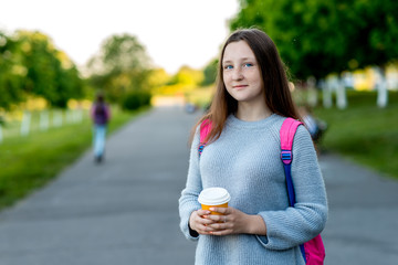 Fototapeta premium The girl is a teenager. In summer On the street in the city. Free space for text. He holds a glass of coffee or tea in his hands. The concept of rest after school. Emotions of happiness smiles.