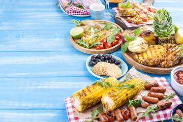 Grilled vegetables, shrimp, fruit on a wooden plate and sausages, juice and salad on a blue background. Summer dinner. Free space for text. Copy space.