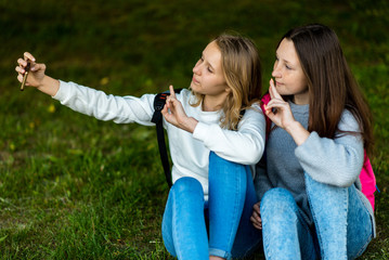 Two teenage girls. In the summer of city park. They sit on the grass and take photos on the smartphone. Behind backpacks. Make a video call over the Internet. Communicates in social networks.