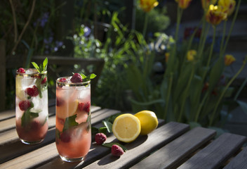 Raspberry ice tea in tall glass with fresh raspberries and lemon. Served on wooden table in a flower garden