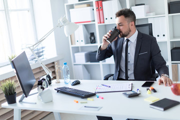 A man sits in the office at the table, drinks coffee and looks at the monitor.