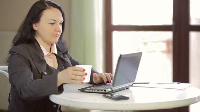Young woman at a table in a cafe, working behind a laptop and drinking coffee. Slow mo. The average plan of the camera moves from right to left.