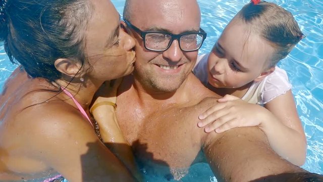 wife and little daughter are kissing man in cheek, bathing in a swimming pool in sunny day