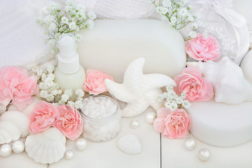 Bridal spa and beauty treatment and cleansing products with carnation flowers, ex foliating salt, seashell soaps, body lotion, sponges, wash cloths with decorative shells and pearls on white wood.