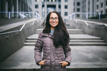 Obraz premium Beautiful young woman of European ethnicity with long brunette hair with a toothy smile, wearing glasses and a coat stands against backdrop of a business center in the fall in cloudy cloudy weather