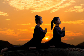 The silhouette of two young girls practicing yoga on the stone at sunset time sky background.