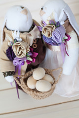 Two handmade rabbits hold a basket with eggs. White wood background. Beige and lilac.