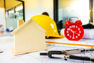 Red clock on archetecture blueprint with pencil, yellow helmet, wooden house model and engineering equipments in modern working office room