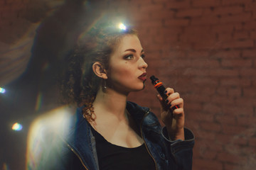 Young attractive red-haired girl smokes an electronic cigarette. She stands by the brick wall. Evening city lights glare.
