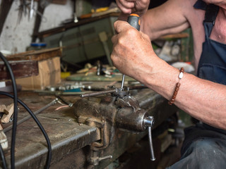 Older man sitting at workbench and working. Human's hands in process of screw cutting, close up view. Home workshop in harness. Hand tools on worktop. Blurred background, soft selective focus