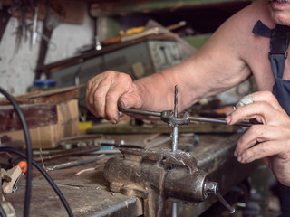 Older man in harnees at home workshop. Close up view on process of screw thread turning. Worker is sitting at home workbench and making thread cutting. Blurred background, soft selective focus