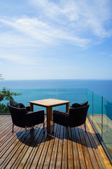 Table and rattan outdoor armchair setting on wooden deck and glass balcony with panoramic Andaman ocean sea view, green tree and blue sky background