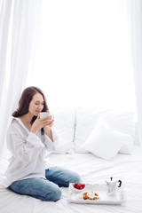Caucasian young woman sitting on white bed at morning. smelling coffee. breakfast, wafers, strawberry