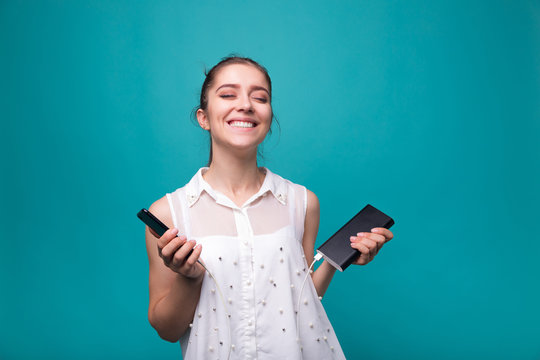 Girl with phone and power bank in hands Young beautiful woman smiles with a phone and charging in his hand blue cyan background 