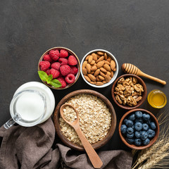 Healthy breakfast ingredients: oat flakes, blueberry, raspberry, honey, walnuts, almonds and almond milk on dark grey concrete background. Square crop. Healthy lifestyle concept