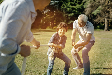 Determined to win. Happy smiling vigorous boy playing rope-pulling with his dad and bending down...