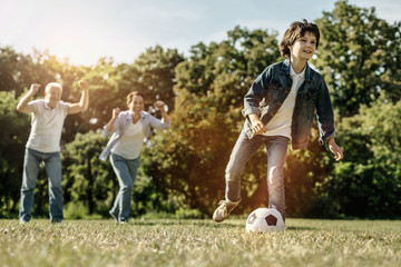 Obraz na płótnie Canvas Feeling excited. Young smiling running boy playing football in nature and his dad and grandpa holding their hands up and supporting him while the boy hitting the ball