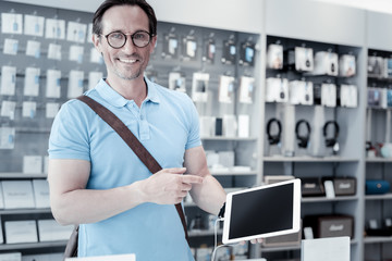 Huge device. Pleasant kind smiling man standing in a new shop and showing a big modern tablet