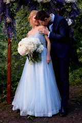 The bride and groom stand at the arch, decorated with flowers, with a large bouquet of white peonies. On-site registration in the Park Wedding walk and photo shoot