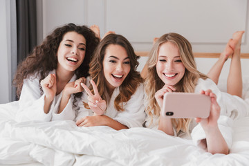 Three joyful girls 20s wearing white bathrobe lying in fashionable bedroom in hotel room, and...