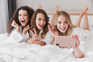 Three european adorable women 20s wearing white bathrobe lying in luxury bedroom at home, and...