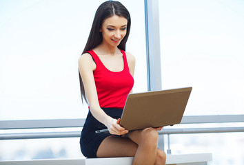 Business. Young Business Woman Sitting at The Table and Works by Computer. Girl Dressed in Red T-Shirt and Black Skirt. The Girl Smiles.