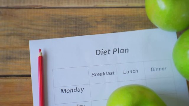 Diet plan. Camera moves and removes from above.Ripe green apples, blank empty diet plan and pencil lie on a wooden table.