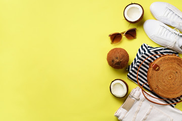 Obraz na płótnie Canvas Female white sneakers, jeans, striped t-shirt, rattan bag, coconut and sunglasses on yellow background with copy space. Top view. Summer fashion, capsule wardrobe concept. Creative flat lay.