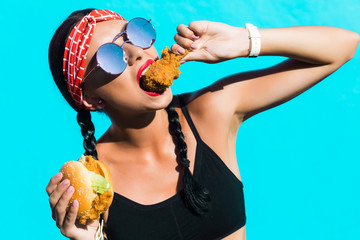 sporty stylish girl in tights, a black top, a bandage on hair, stands on the background of a colored wall. He eats a burger and a piece of fried chicken, drinks soda. Strong emotions, smile, fast food