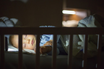 Adorable newborn baby boy, sleeping in crib at night. Little boy in white pajamas taking a nap in dark room. Bedroom interi?r with lamp and teddy bear . © Tomsickova