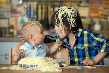 Little baby boy and his older brother, toddler child, eating spaghetti for lunch and making a mess