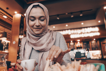 beautiful smiling Arab girl in hijab having lunch in cozy cafe, drinking fragrant tea