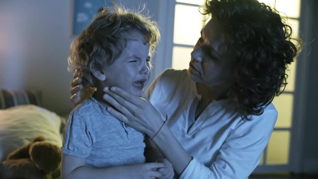 Medium shot of worried mother kissing and hugging crying son of primary school age when trying to calm him down at night