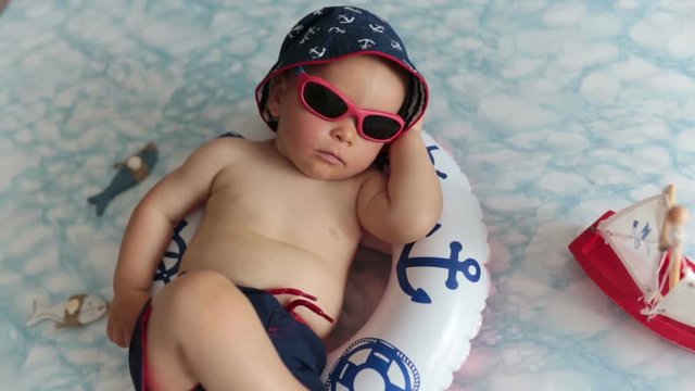 Cute toddler, baby boy sleeping on a tiny inflatable swim ring, wearing swimsuit shorts and sunglasses, indoor shot