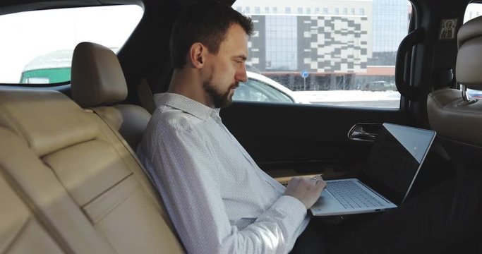 Young businessman with a beard uses a touchpad on a laptop on backseat of car