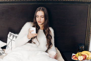 long-haired beautiful girl lies in bed in the evening, looks into her smartphone