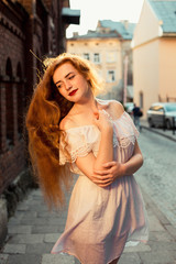 Elegant red haired model with long curly hair wearing white dress posing in evening sunlight