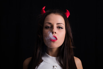 Young woman with Devil horns on head and cigarette smoke posing on black background