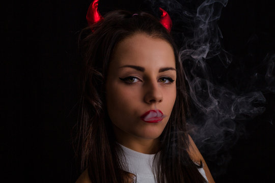 Young woman with Devil horns on head and cigarette smoke posing on black background