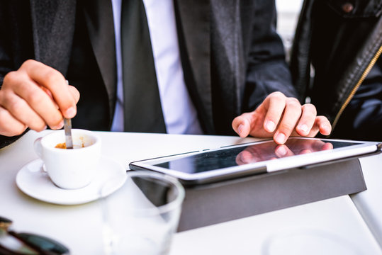 Close up on the hands of young businessman stirring with a tablespoon a cup of coffee and using tablet, tapping screen - technology, work, break concept