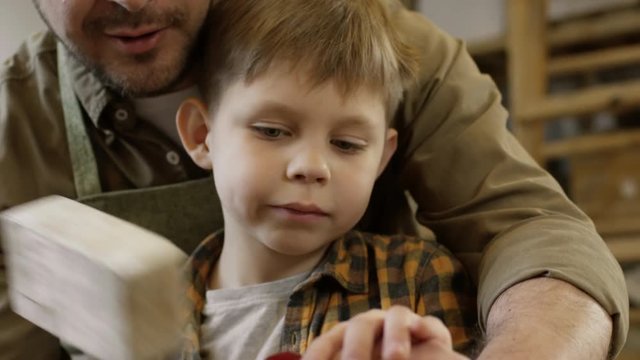 Tilt down of bearded male carpenter teaching little son how to carve wood: he helping him to use chisel and mallet and explaining how to do it correctly