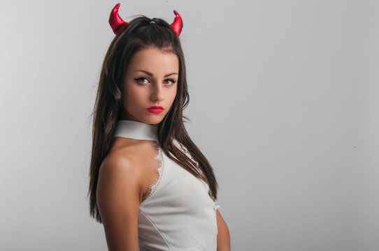 Studio Portrait Of Beauty Brunette Woman Standing In White T-shirt With Red Evil Horns On Head, White Background